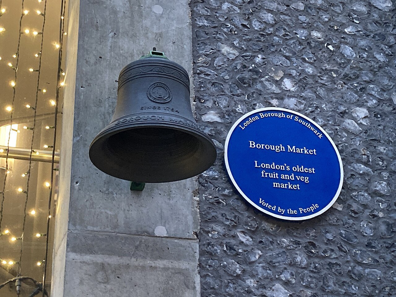 A bronze bell on a wall beside a blue plaque naming Borough Market as London's oldest fruit and veg market