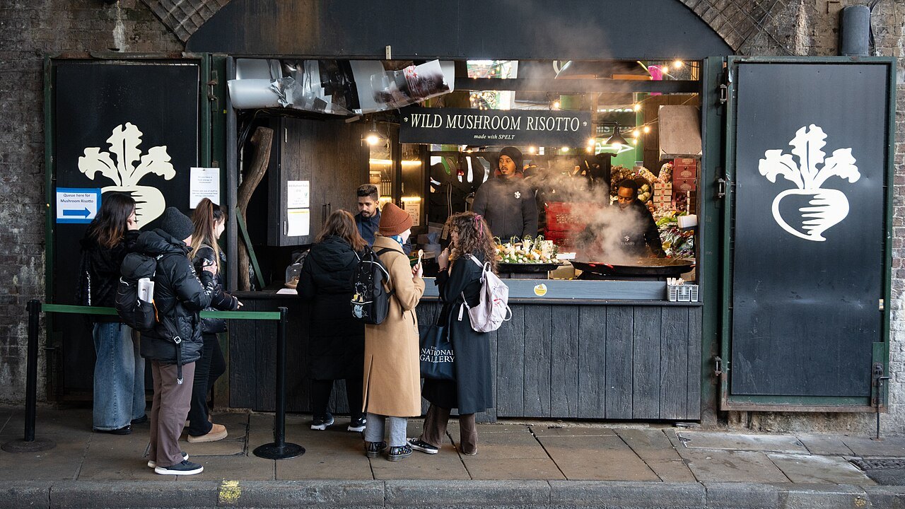 A small market stall under a railway arch, its sign advertising wild mushroom risotto, with customers queuing