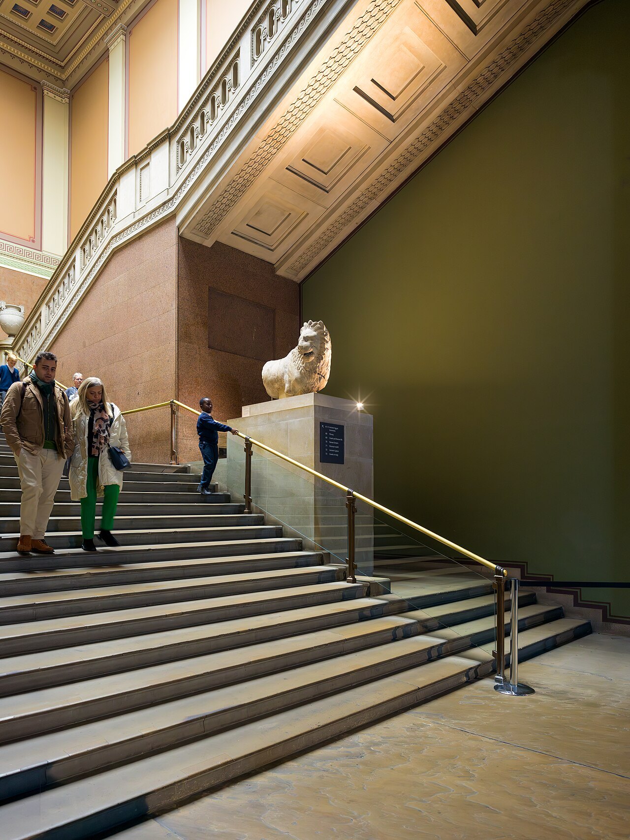 A stone lion beside a stair inside the British Museum, with a handrail along the steps