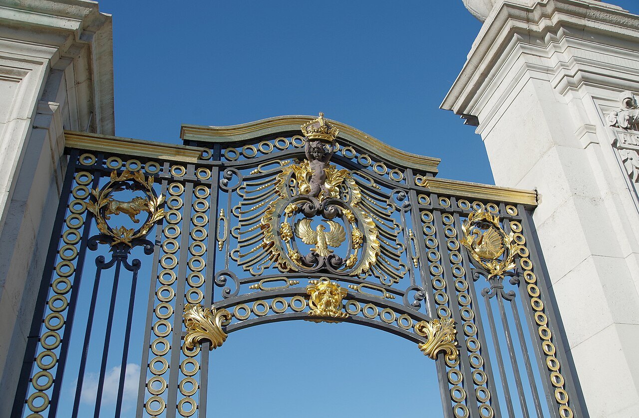 A pair of tall blue and gold gates with a royal crown and scrolling ironwork beside white stone gateposts