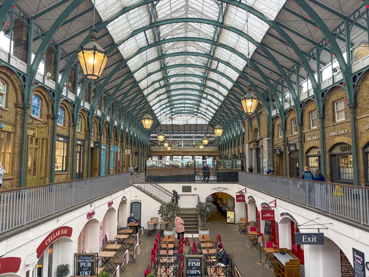 The upper gallery of the Covent Garden Market hall, its arched iron roof lit by lamps, with a restaurant court below