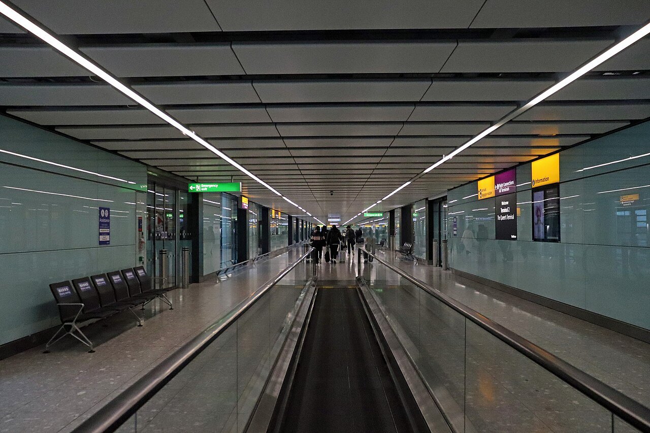 A moving walkway along a glazed corridor at Heathrow Terminal 2, with benches at the side