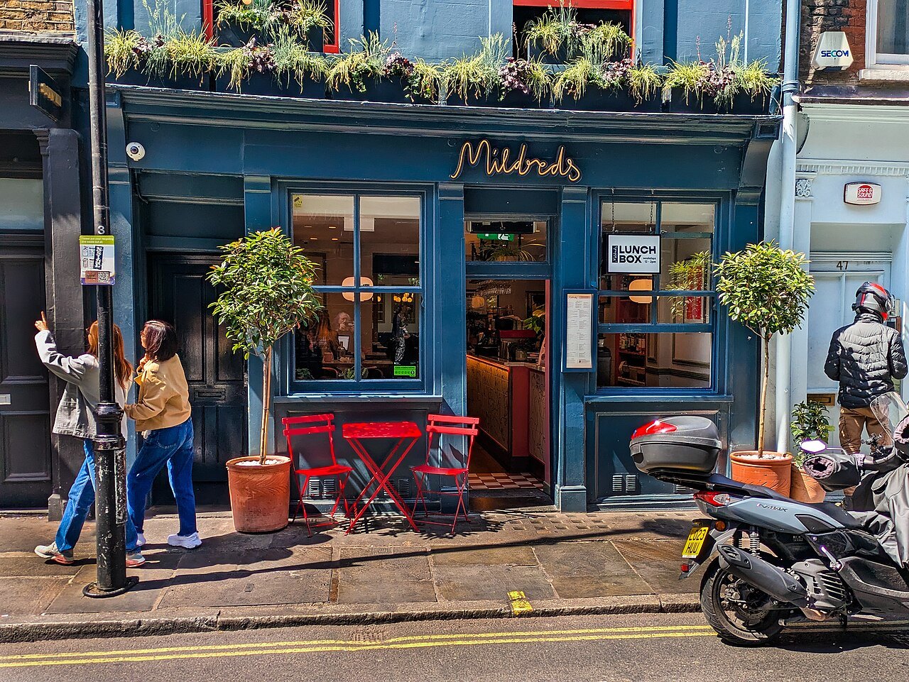 The blue front of a cafe in Soho with red chairs and plants above the window, and people passing on the pavement