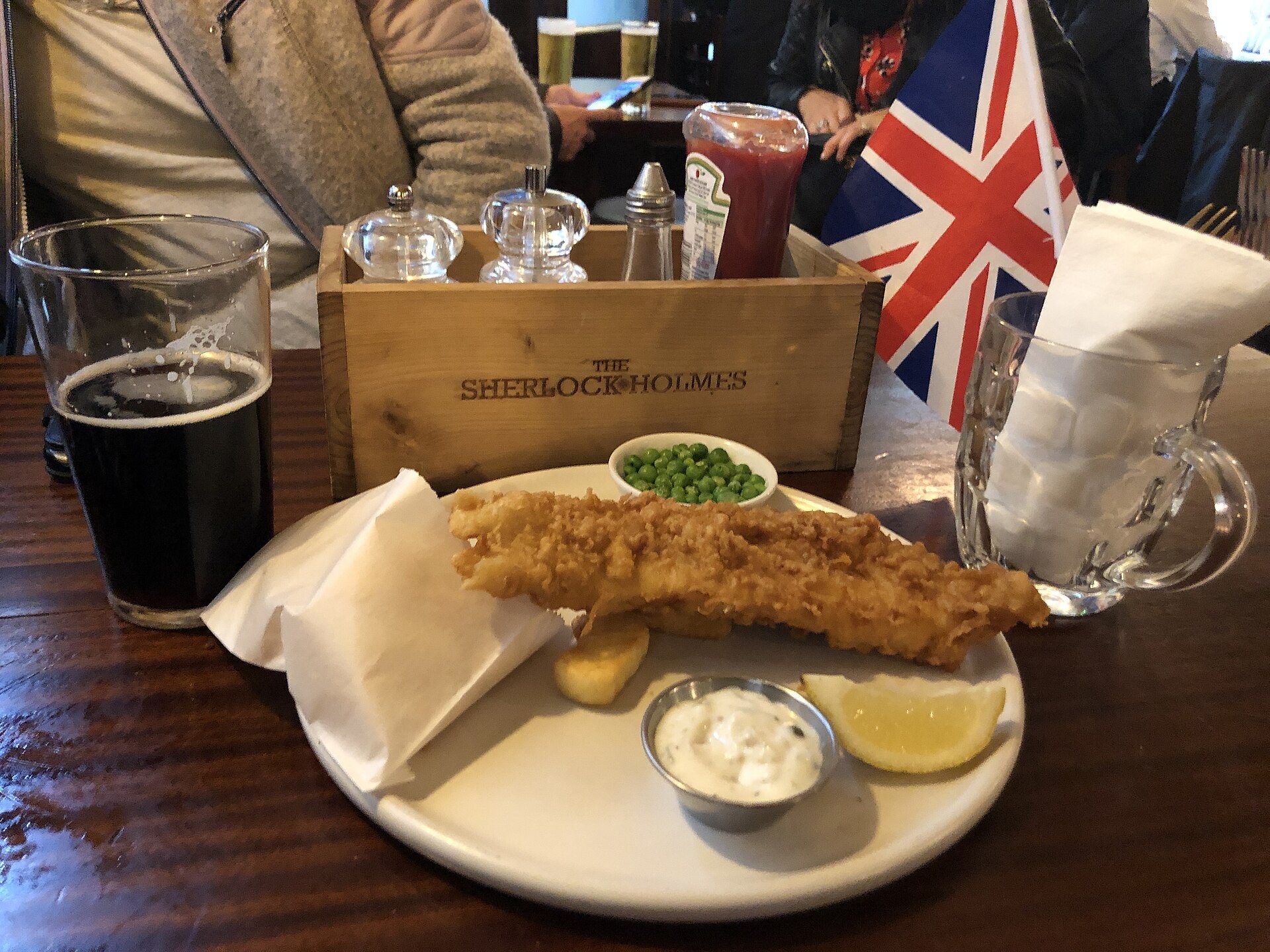 A plate of fish and chips with mushy peas and tartare sauce beside a dark pint and a Union flag