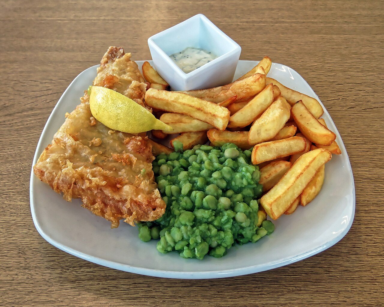 Battered fish, chips and peas on a white plate with a wedge of lemon and a pot of tartare sauce
