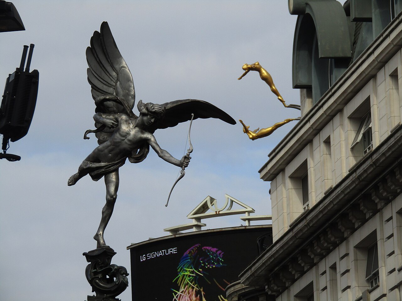 The winged bronze figure of the Eros statue at Piccadilly Circus, with a lit screen behind it