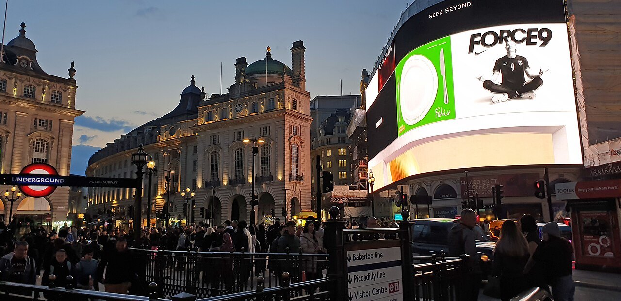 Piccadilly Circus at dusk, with the lit screens at the corner and a crowd on the pavement