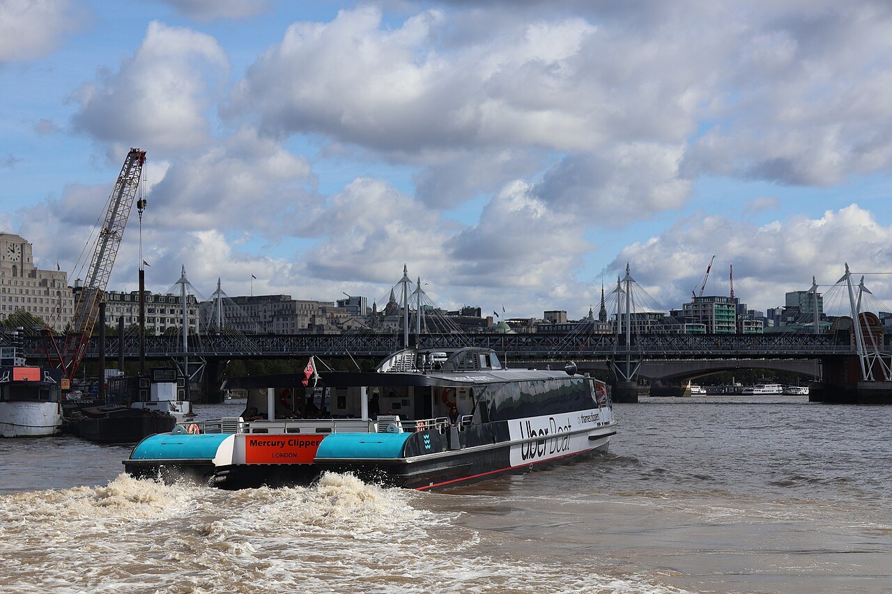 A river bus churning along the Thames at Westminster, with the Hungerford footbridges behind
