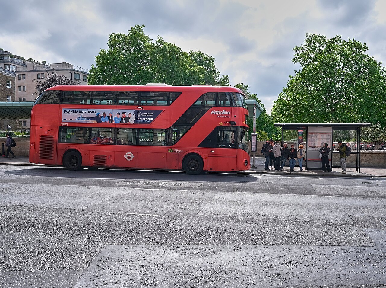 A red double-decker bus pulling out from a stop on a wide road under trees