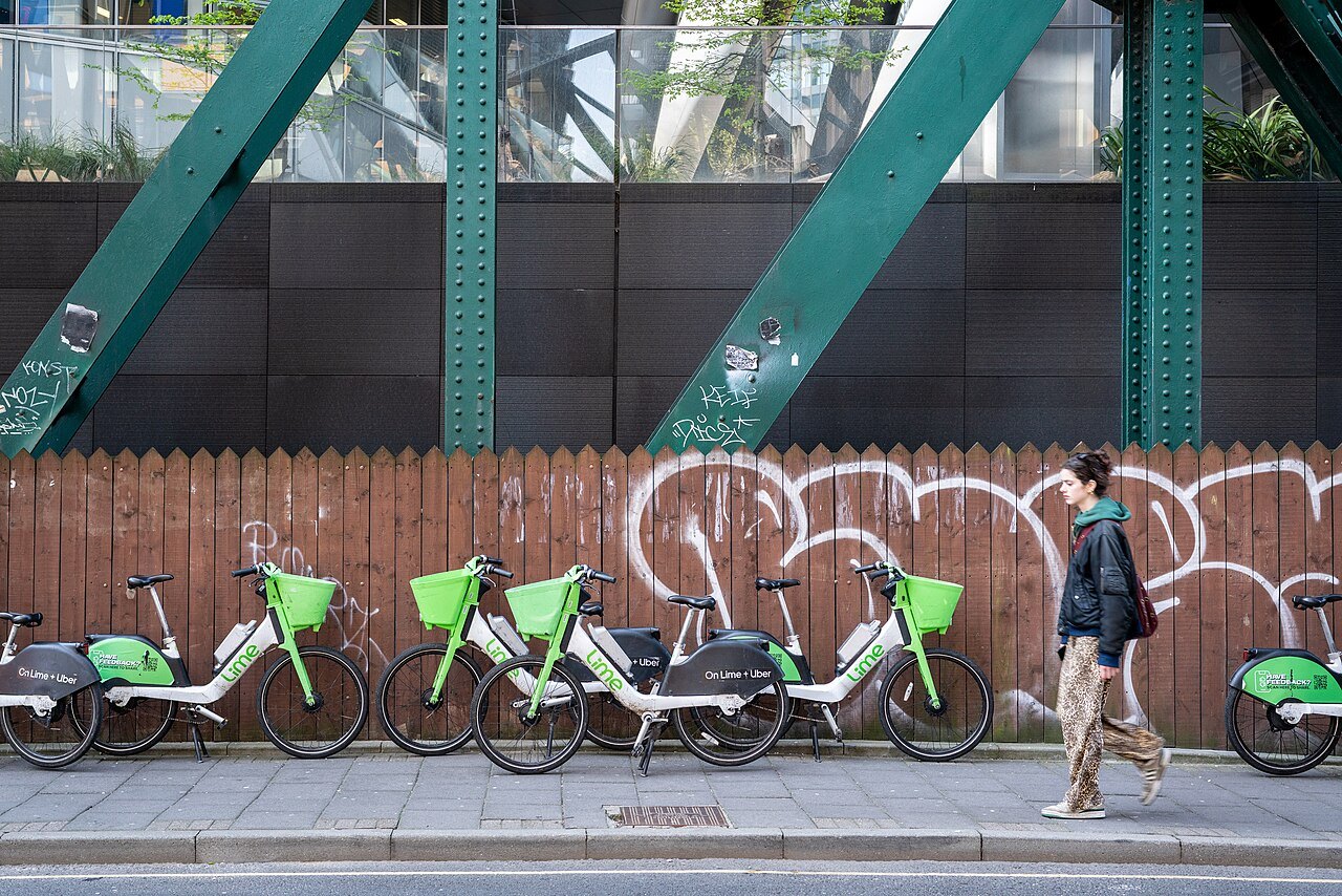 A row of hire bicycles parked on a pavement beside a wooden fence covered in graffiti