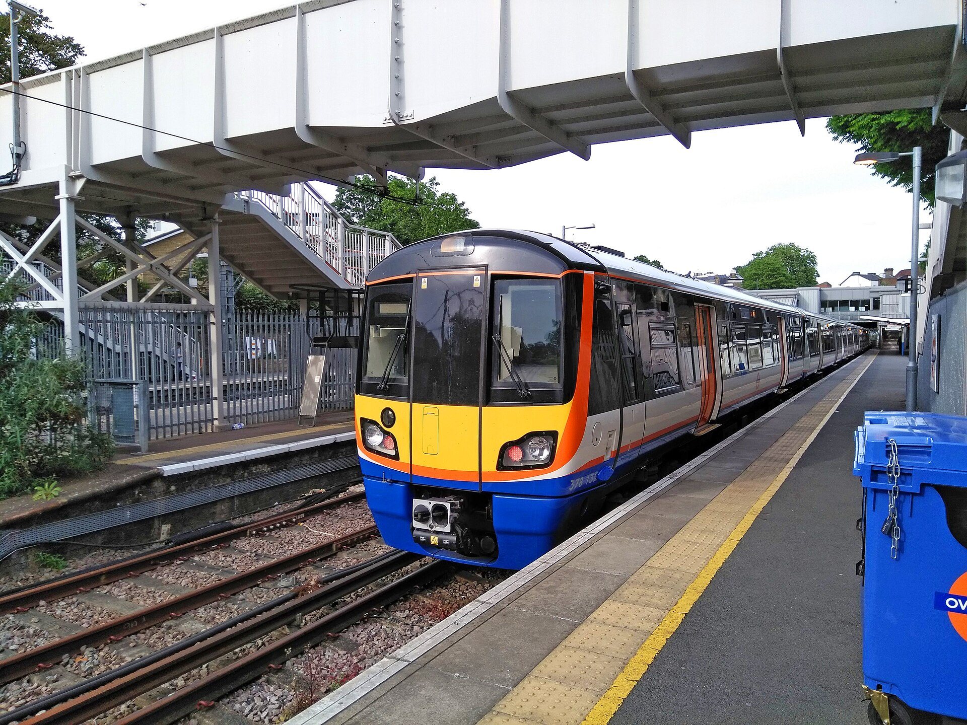 A train in the orange, blue and white colors of the Overground at a platform on a bright day
