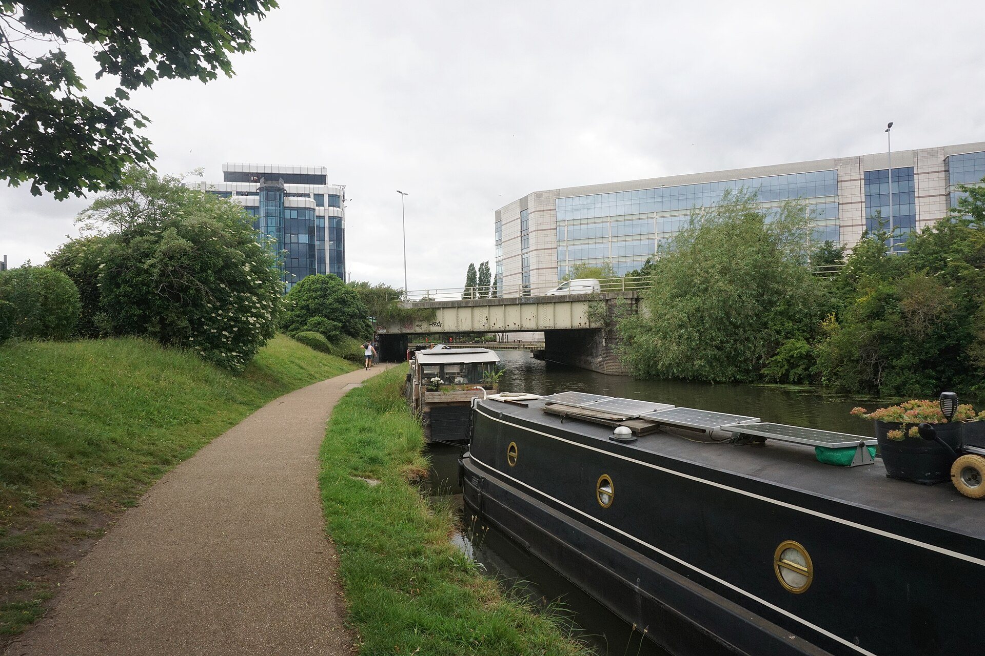 A towpath beside a canal, with a black narrowboat moored at the right and office blocks beyond