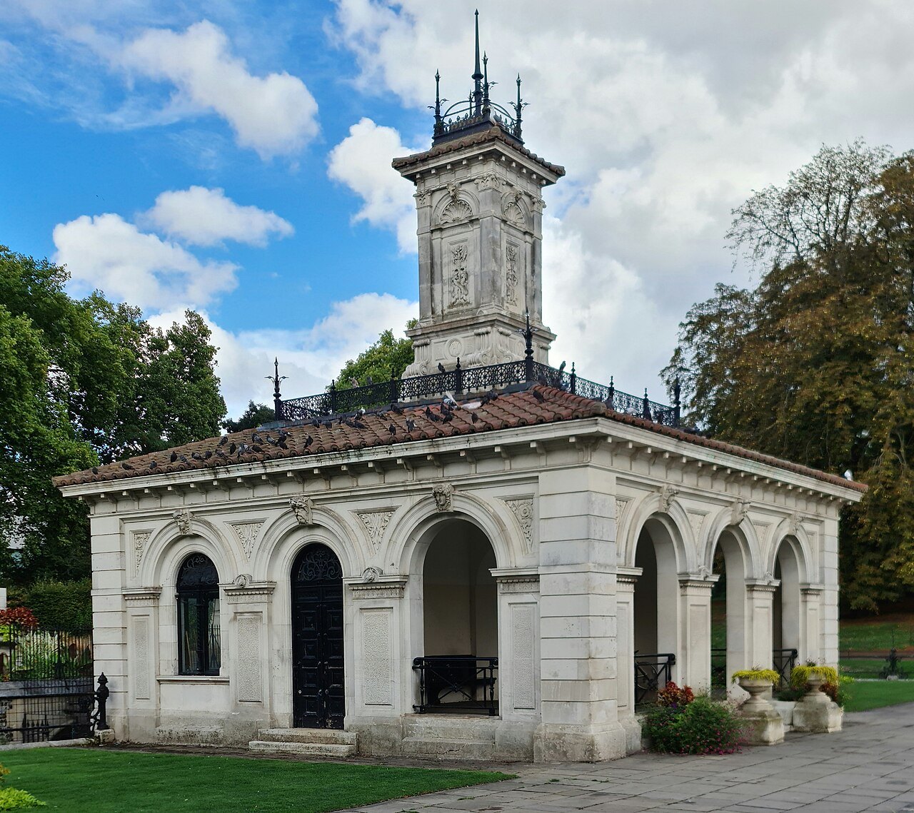 A small stone pavilion with a clock tower and arched openings in Kensington Gardens