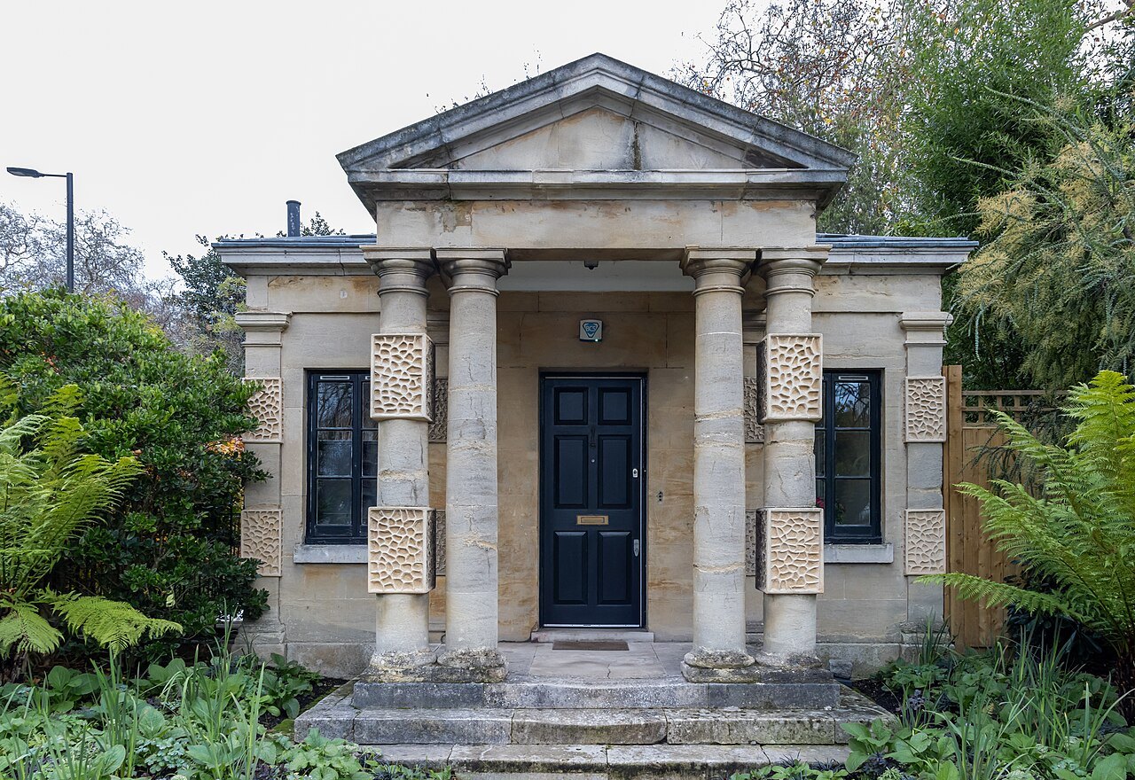 A small stone lodge with four columns and a pediment, beside a gate in Hyde Park among ferns and trees