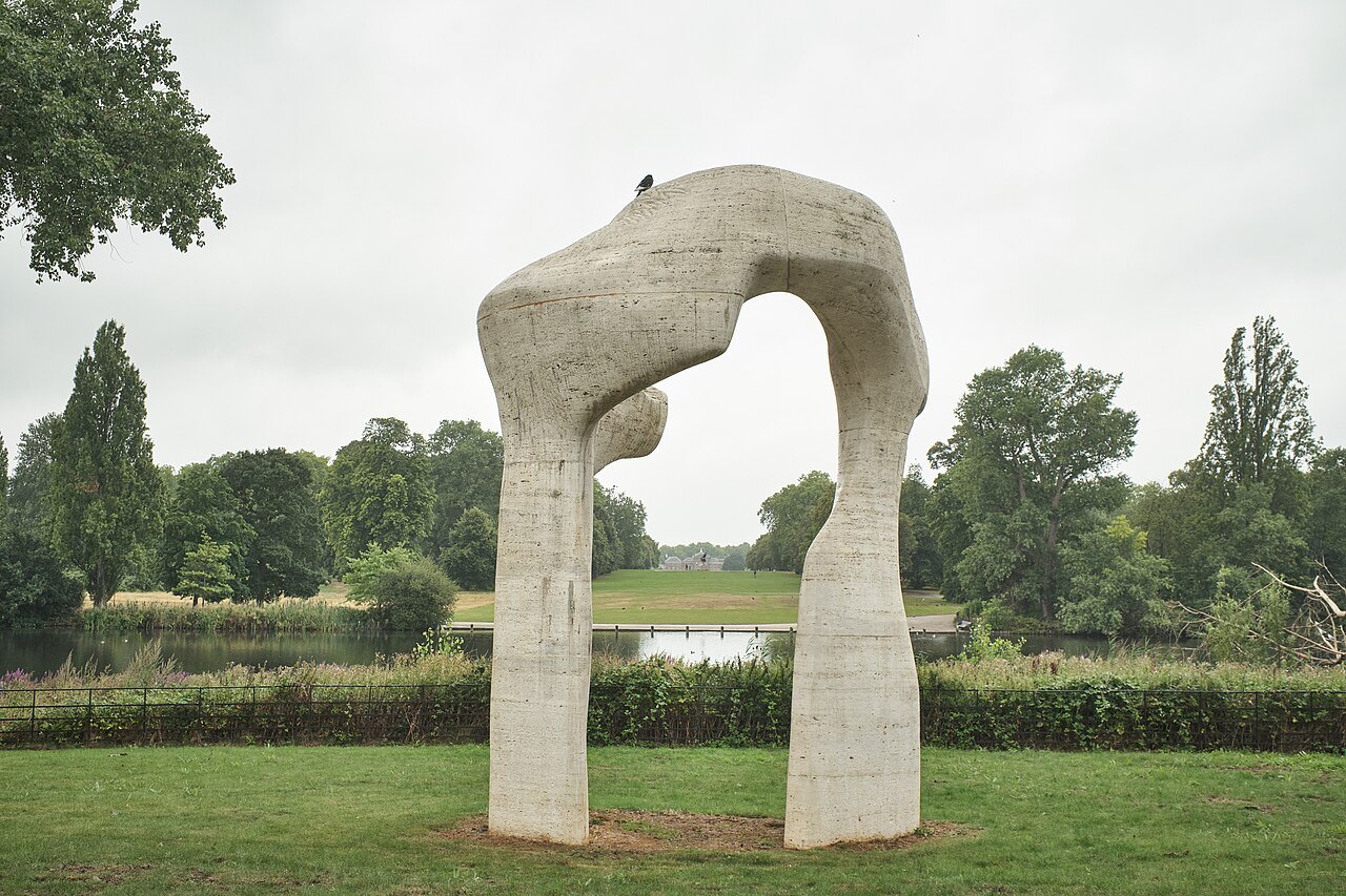 A tall stone arch sculpture standing on the grass beside a lake in a park