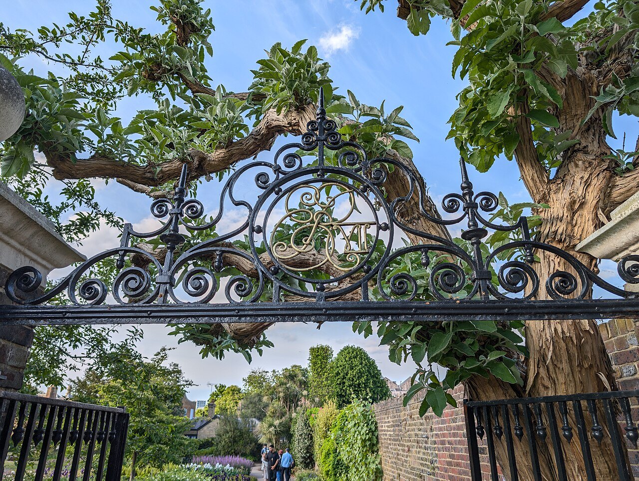 The iron gate of the Duke's Garden at Kew, scrolled and crowned, framed by leafy branches