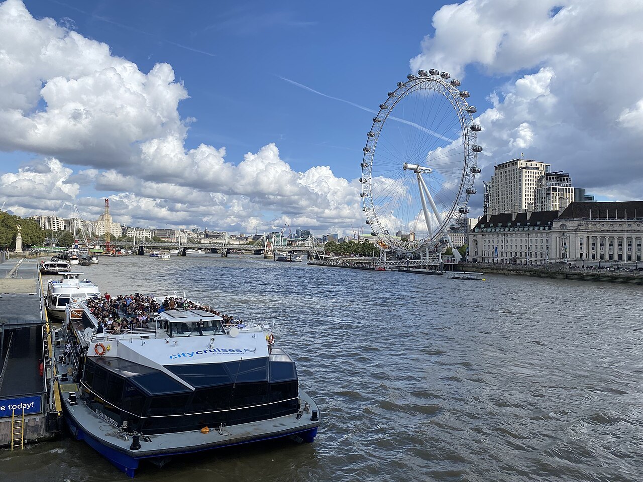 A river boat crowded with passengers at a Thames pier below the London Eye