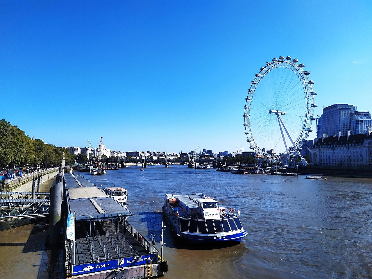 A river boat at a Thames pier with the London Eye rising behind it under a deep blue sky