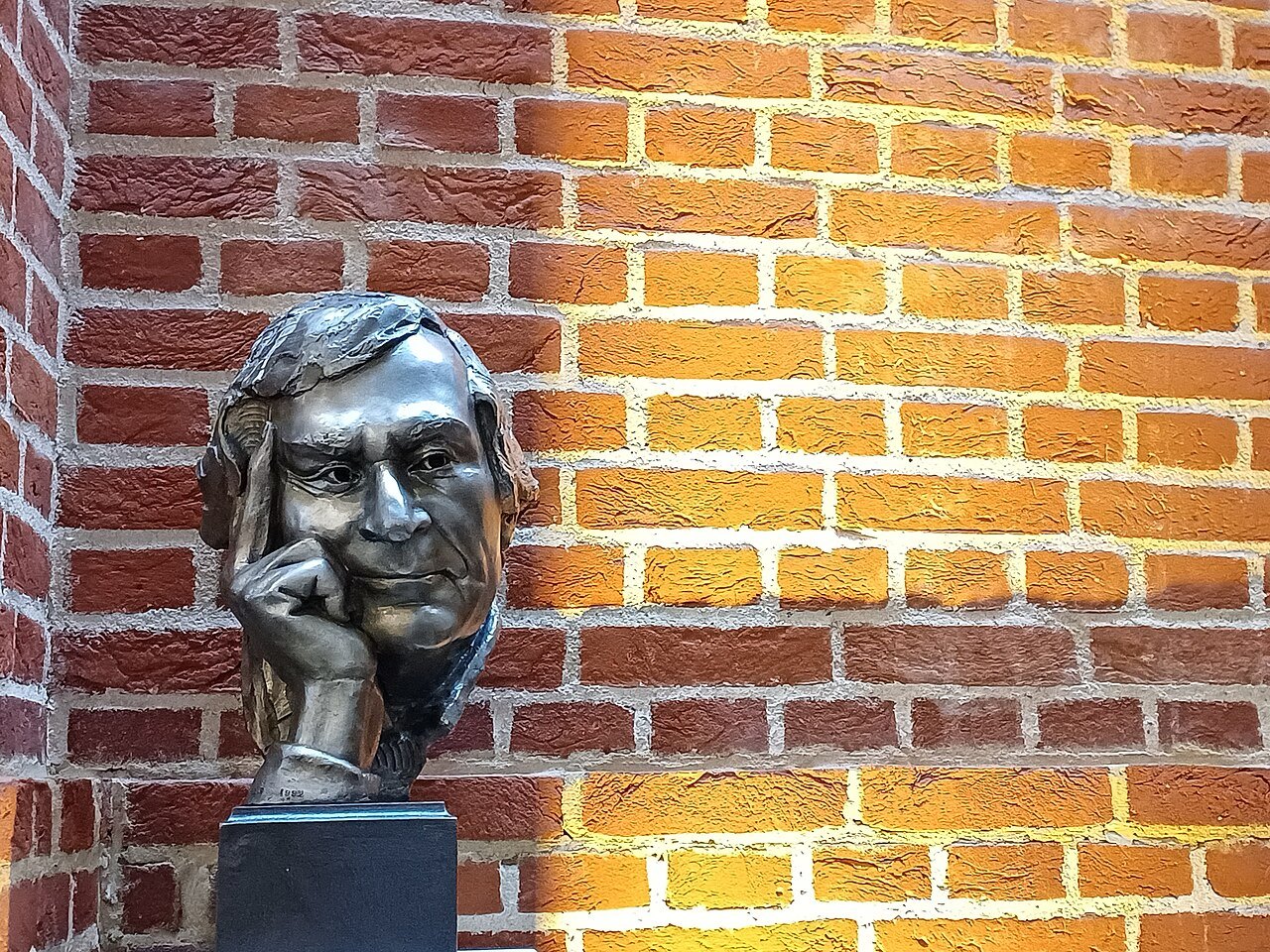 A bronze head of Sam Wanamaker against a wall of red bricks, in sunlight