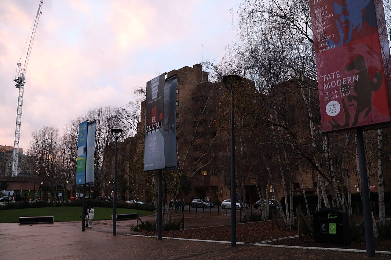 The brick side of Tate Modern seen through bare birch trees, with a banner advertising an exhibition