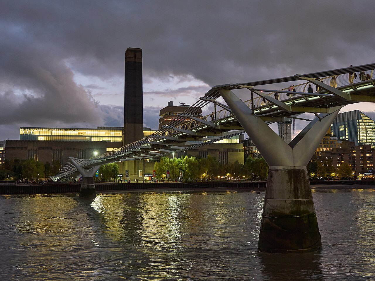 The Millennium Bridge and the Tate Modern chimney at dusk, the bridge's lamps reflected in the river