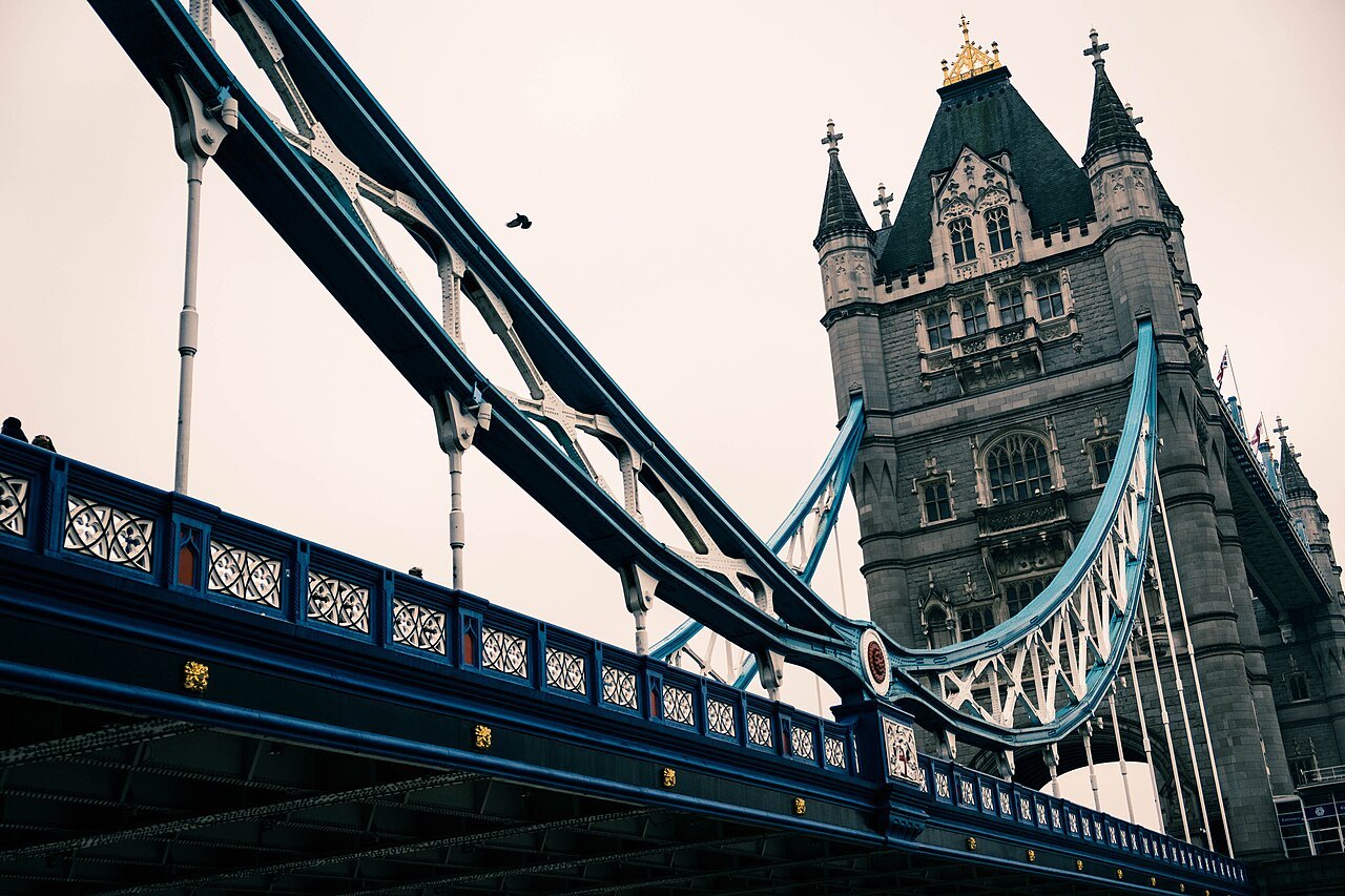 A tower of Tower Bridge and its blue girders seen from the roadway, looking up at the walkway