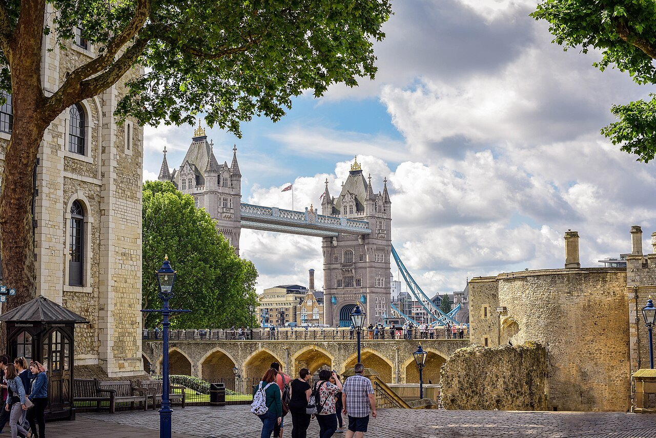 Tower Bridge seen through the trees from inside the Tower of London, with visitors on the paved terrace