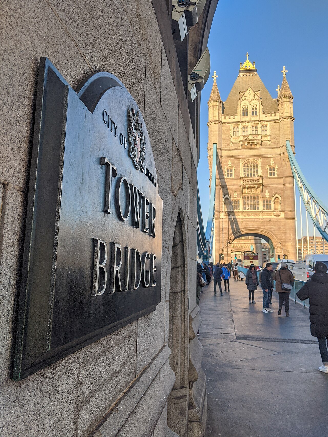 A dark plaque reading Tower Bridge on a wall, with the bridge's tower and walkers beyond