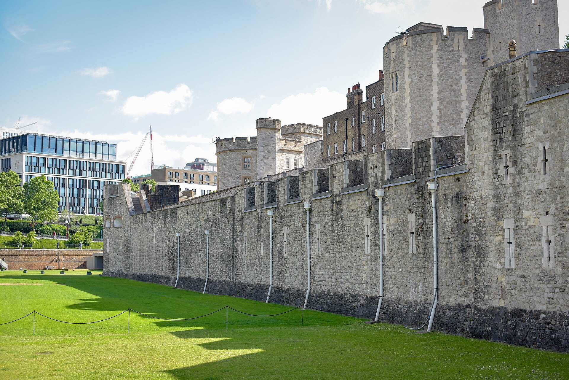 A long stretch of the Tower of London's outer wall above its dry moat, the grass in sunlight