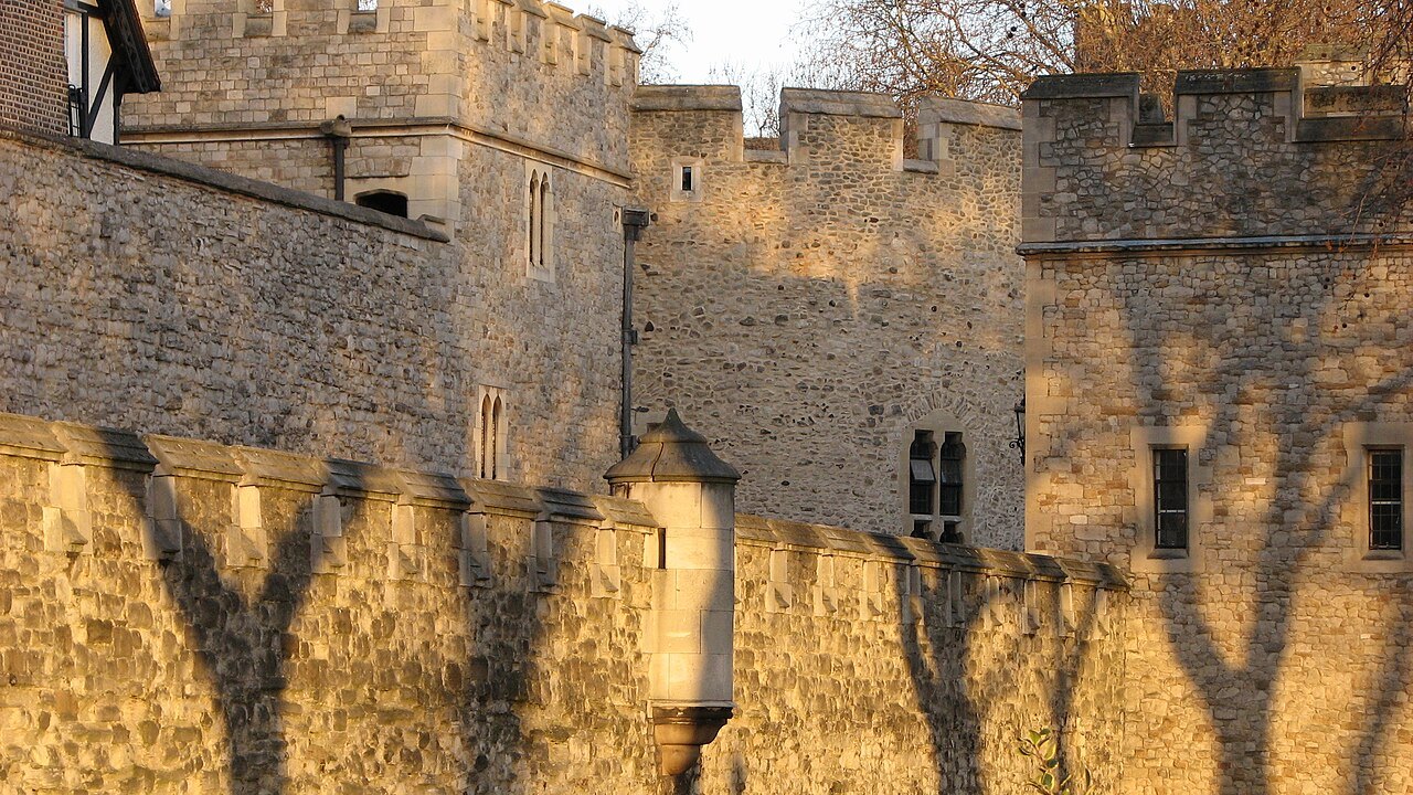 The Tower of London's walls in low sunset light, with the shadows of trees cast across the stone