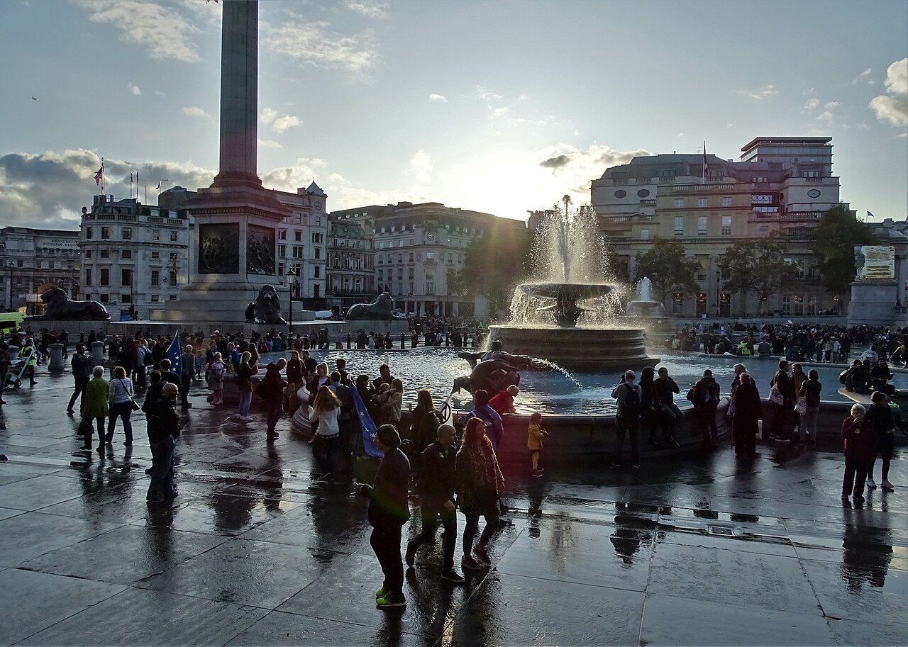 Trafalgar Square in backlight at the end of a wet day, with Nelson's Column and the fountain beyond the crowd