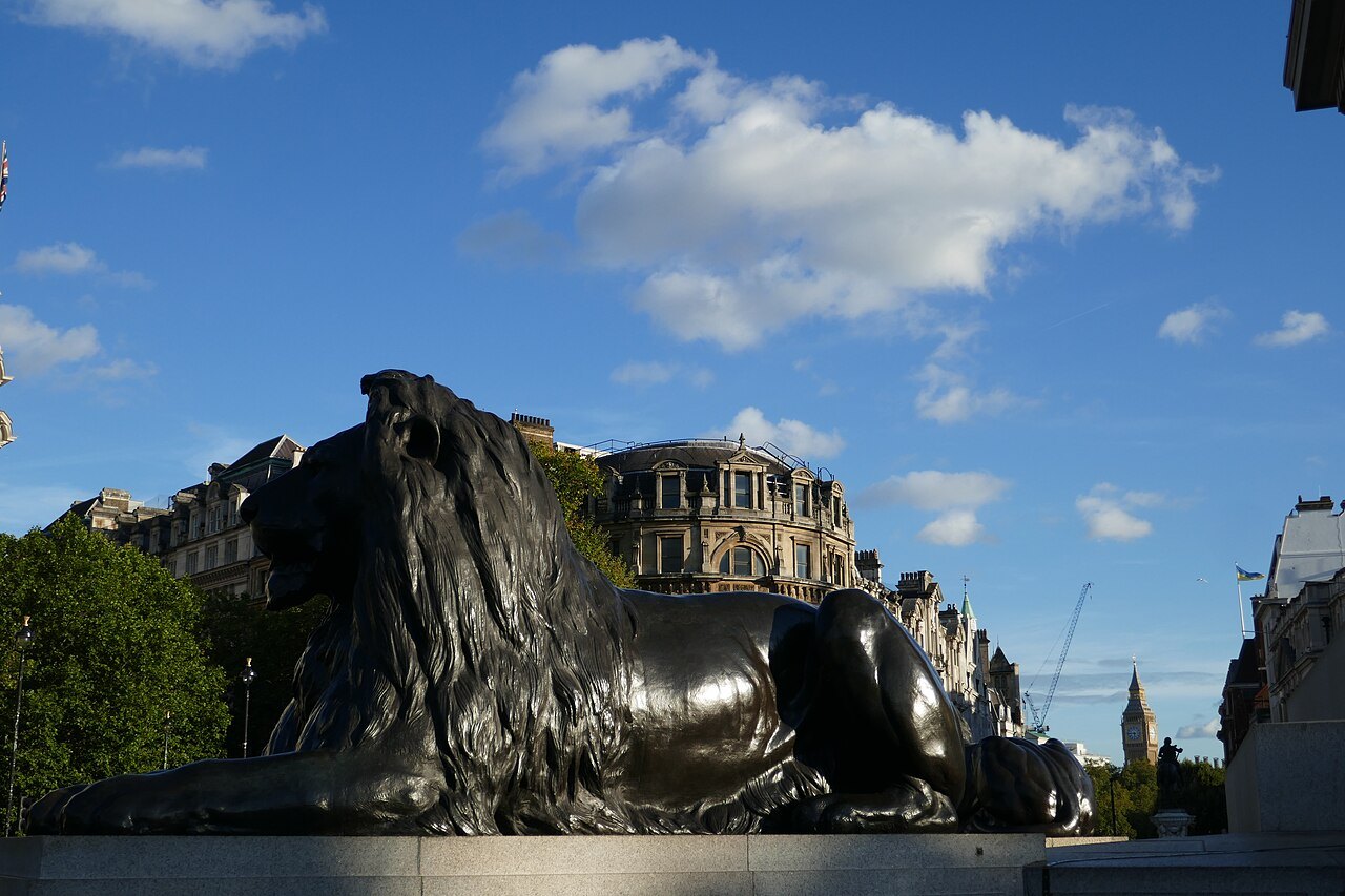 A bronze lion lying in the sun beside the column's base, with trees and buildings behind