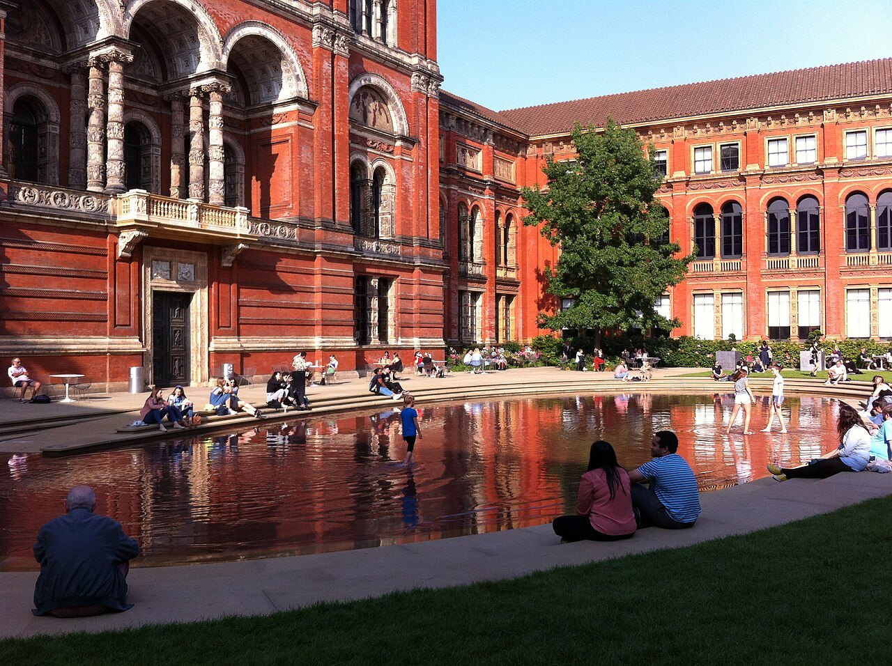 Visitors sitting at the edge of the shallow pool in the museum's courtyard, in front of the red brick arches