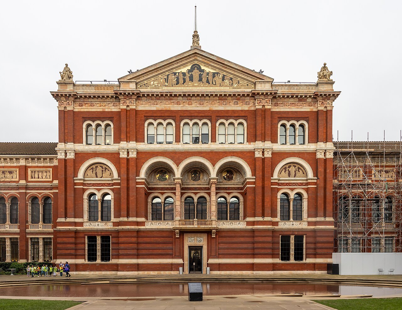 The red brick and terracotta front of a V&A building above its garden, with scaffolding at the right