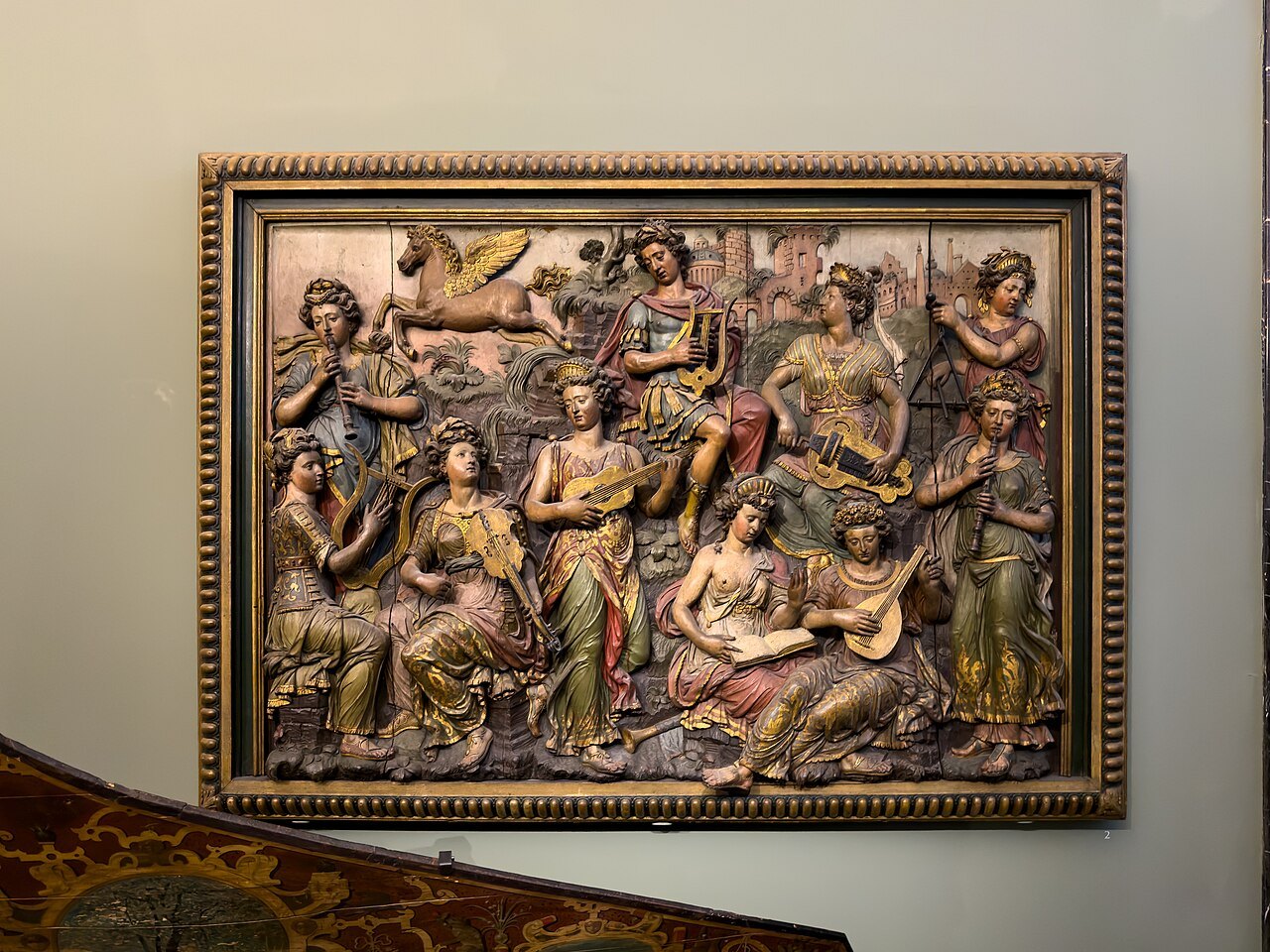 A painted and gilded relief of figures playing instruments, in a carved frame on a gallery wall at the V&A
