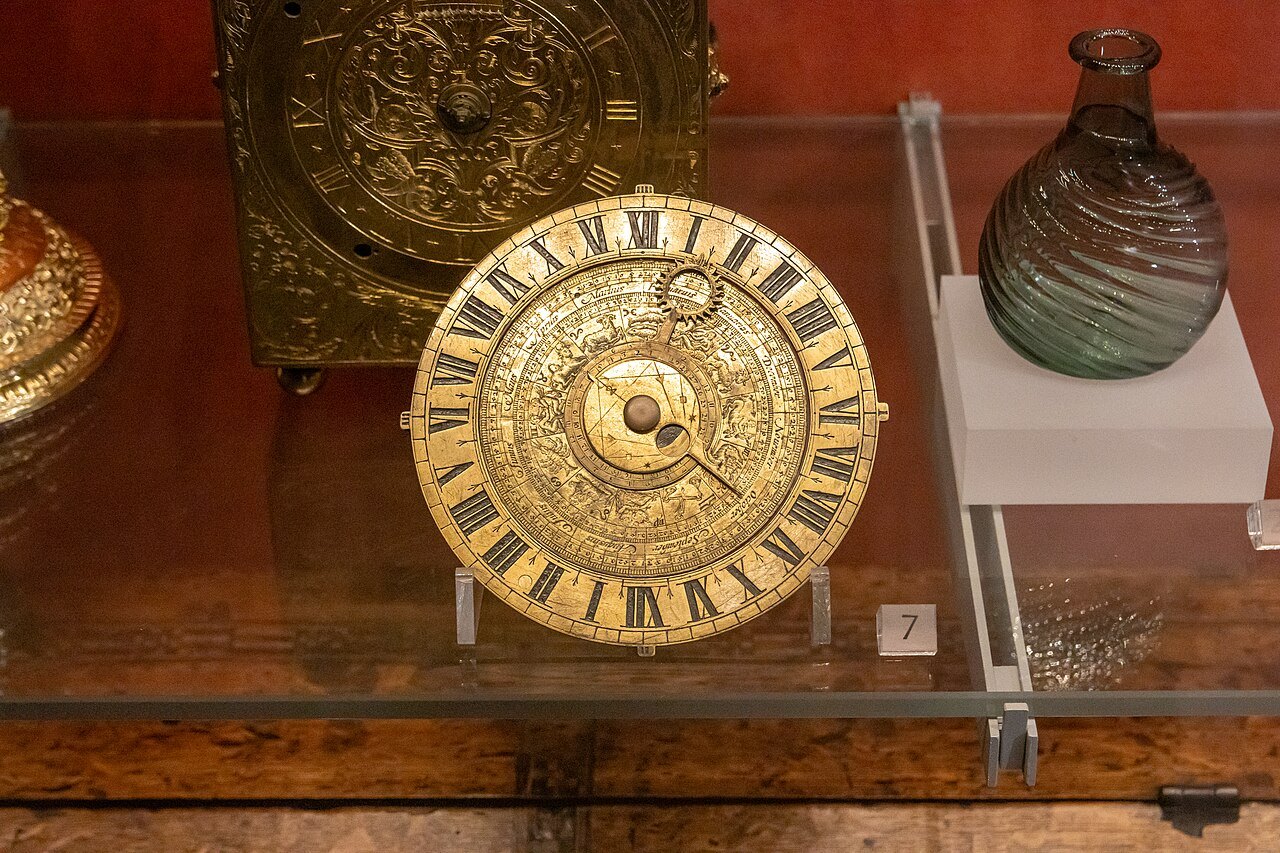 A gilt table clock with Roman numerals in a glass case beside a glass flask at the V&A