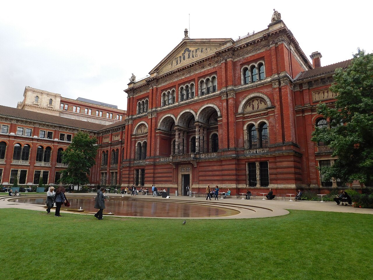 A walkway round the pool in the V&A courtyard, with the brick front of the museum behind and the lawn in front